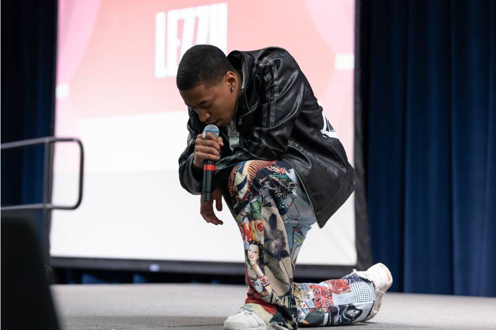 Lezzu kneeling on stage singing into wireless microphone.